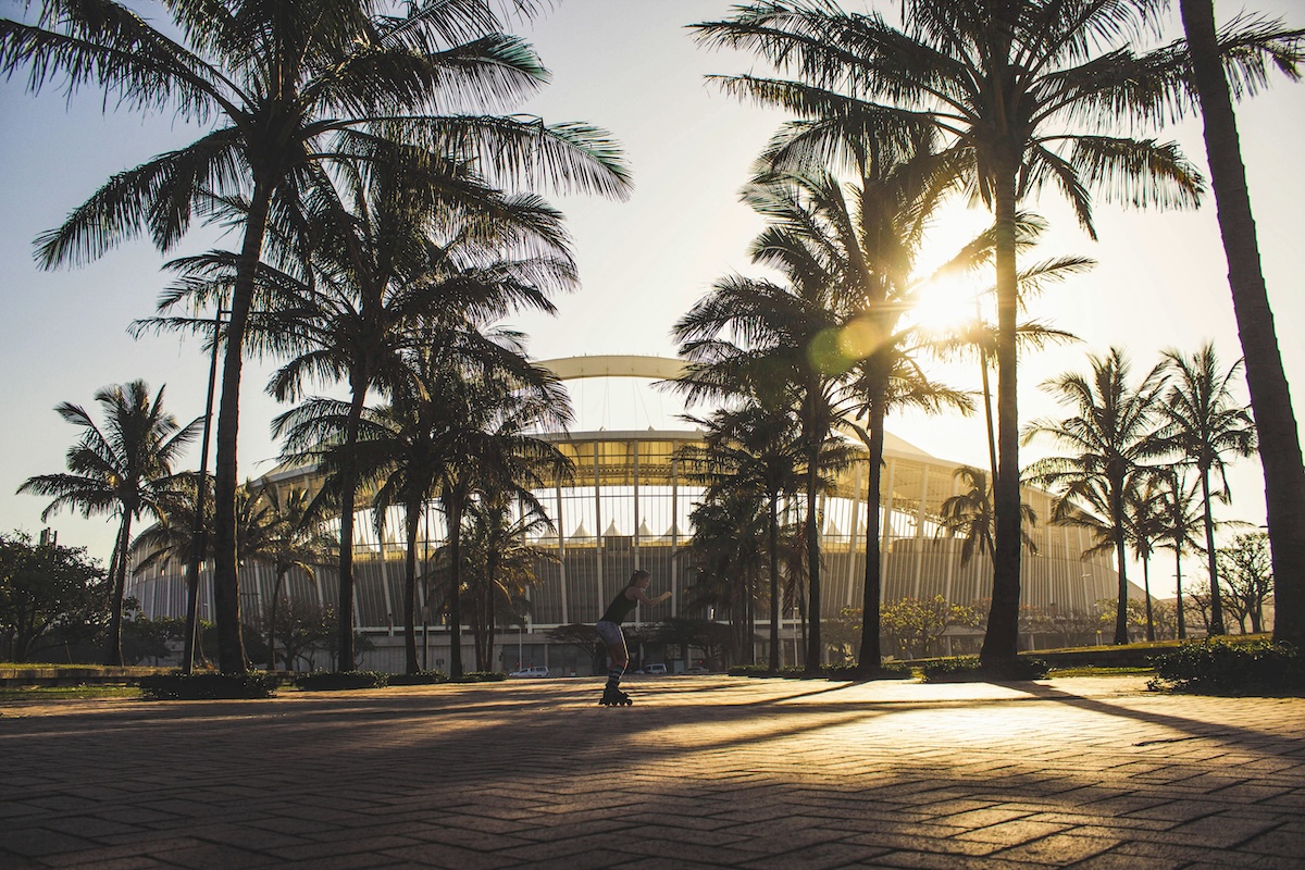 A picturesque view of Durban with palm trees silhouetted against the sunset, framing the iconic Moses Mabhida Stadium.