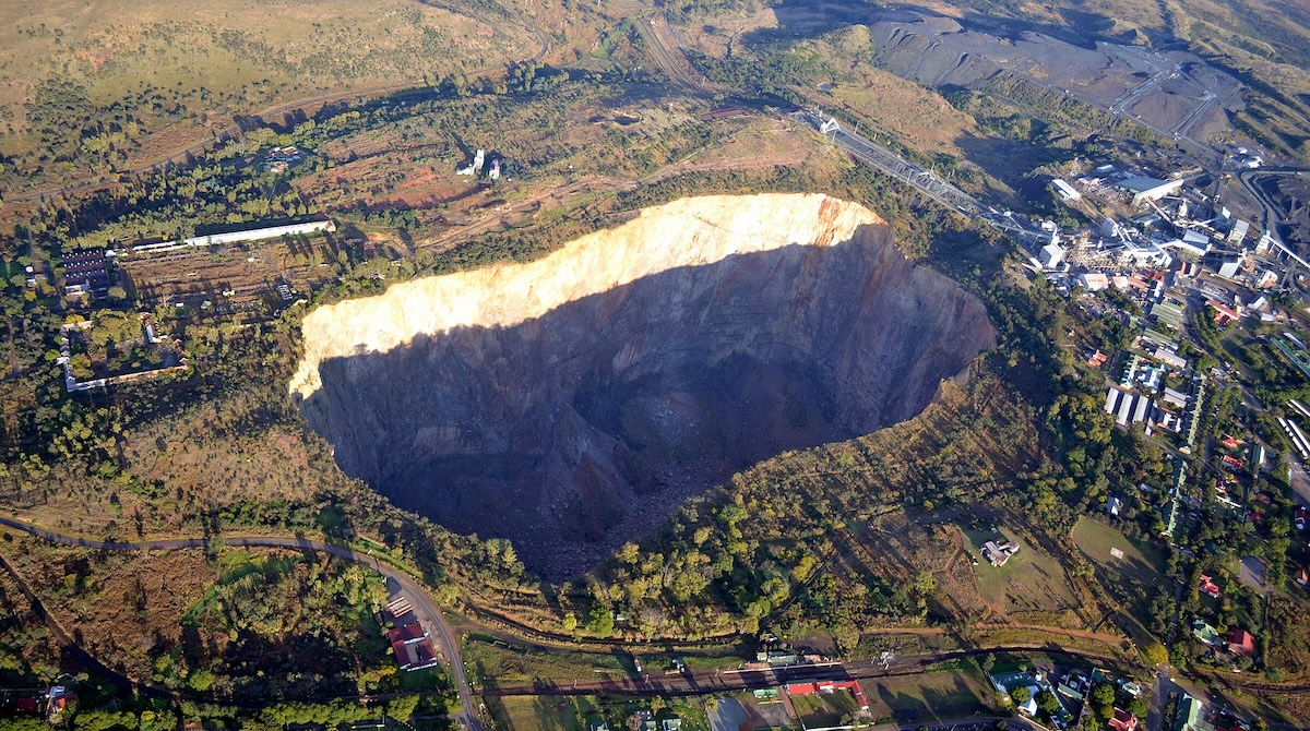 An aerial view of a large open-pit mine in South Africa, surrounded by lush green landscape and industrial buildings.