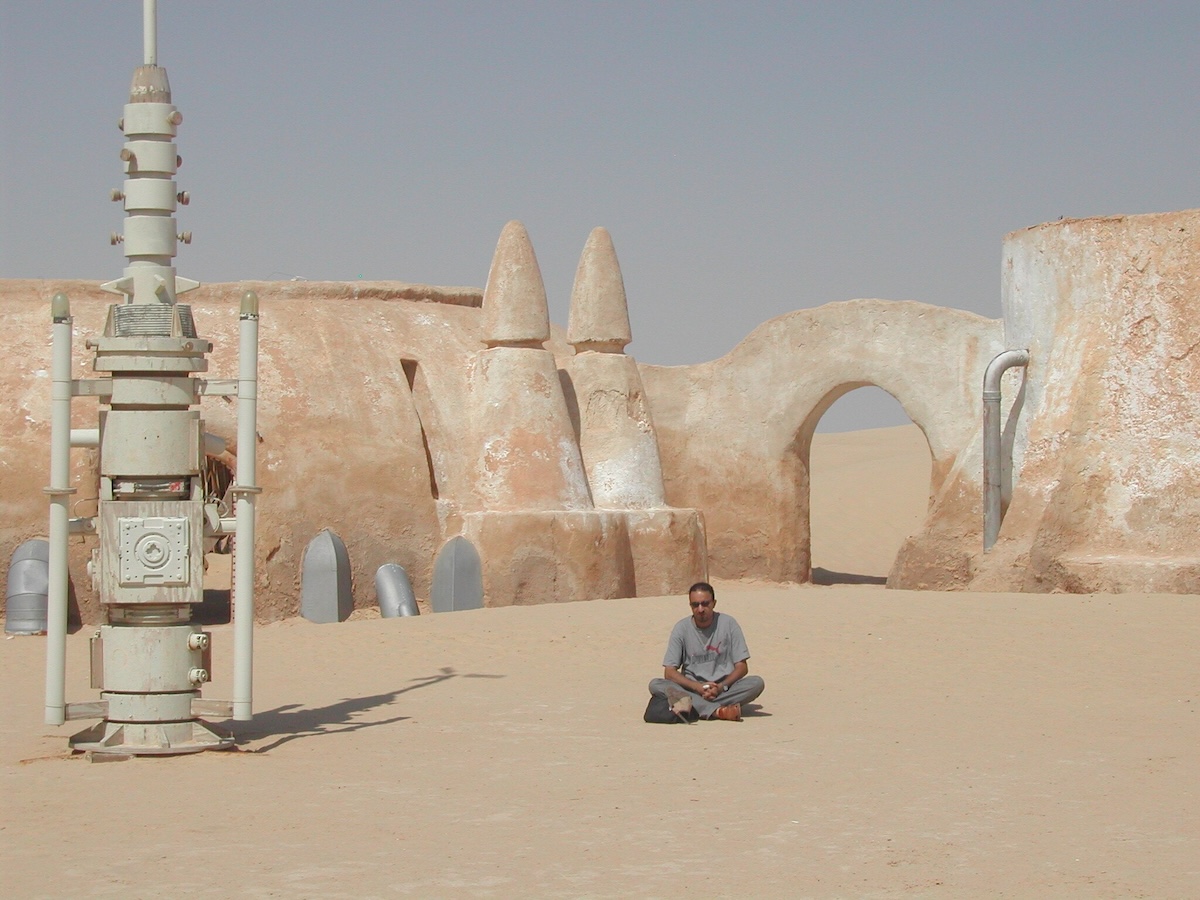 A photograph of a person sitting in front of the Star Wars film set in the Tunisian desert, featuring iconic Tatooine architecture.