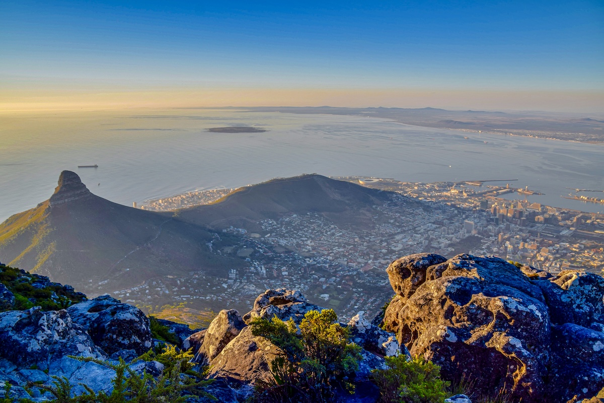 An elevated view from Table Mountain, overlooking the city of Cape Town and the surrounding coastline, under a clear blue sky at sunset.