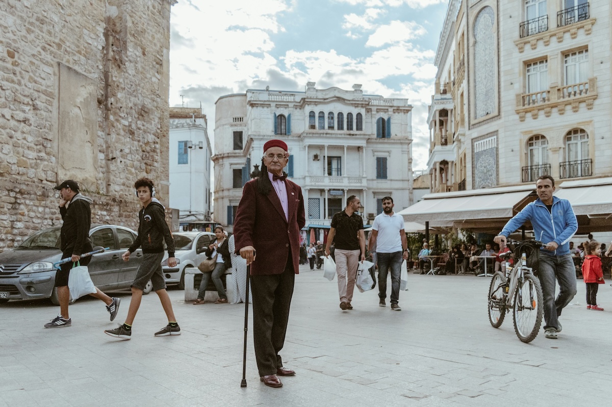 A street scene in Tunis with a blend of historical and modern buildings, people walking, and a man in traditional attire in the foreground.