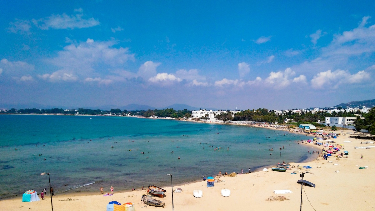 A bustling beach scene in Tunisia with people swimming and sunbathing, and a backdrop of turquoise waters and a clear sky.